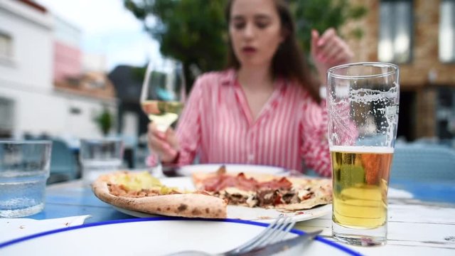 Young pretty woman eats pizza and drinks white wine at outdoors restaurant. Freshly baked Italian pizza on table. Beer on foreground. Slow motion.