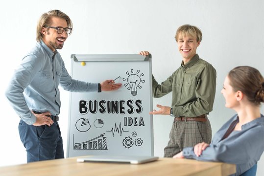 Laughing businessmen showing business idea inscription and icons on flipchat to businesswoman