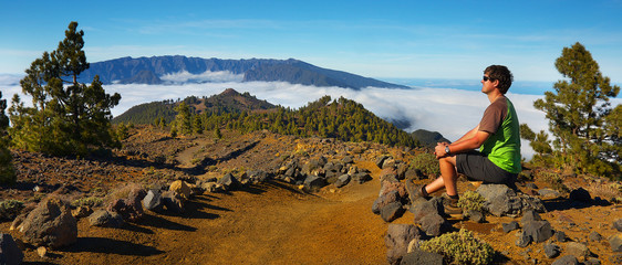 Man sitting on the stone watching a volcanic landscape of pine forest with a Caldera de Taburiente on background, island of La Palma, Canary Islands, Spain