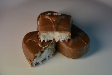 On a white glass plate there are beautiful sweets with coconut filling of white color and milk chocolate, pieces from a bar