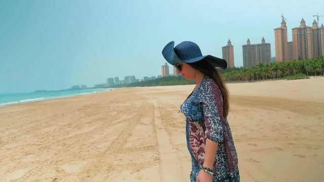 Beautiful happy girl in blue hat walks on beach on sunny summer day against a blue sky. Medium Shot.