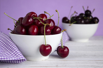 Ripe cherry berries in two white plates on a white wooden background, lilac in a cage towel. Vegetarian concept, food.