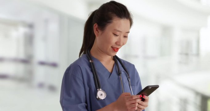 Close Up Of Female Millennial Nurse Dressed In Scrubs Using Smartphone To Send Text Messages Indoors Hospital, Happy Young Asian Doctor Texting With Cell Phone Inside Clinic, 4k