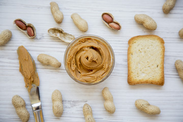 Toast, bowl of peanut butter, knife and peanuts in shells on a white wooden background, top view. From above.