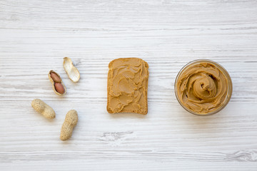 Toast, bowl of peanut butter and peanuts on a white wooden background, top view. Flat lay.