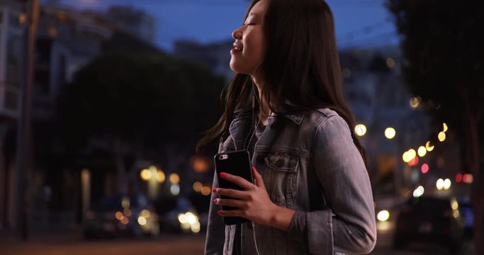 Cool millennial woman using smartphone to listen to music and wearing denim jacket at night in San Francisco, Close up of cute Asian woman dancing to music on urban street, 4k