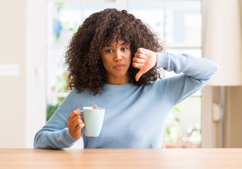 African american woman holding a cup of coffee at home with angry face, negative sign showing dislike with thumbs down, rejection concept