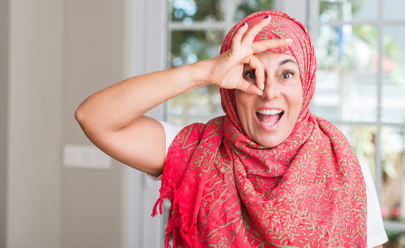 Middle aged muslim woman wearing hijab with happy face smiling doing ok sign with hand on eye looking through fingers