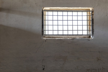 Old white wall with window covered by iron grate. Sun shining  through grill. Prison room interior . Enlightment concept. Copyspace