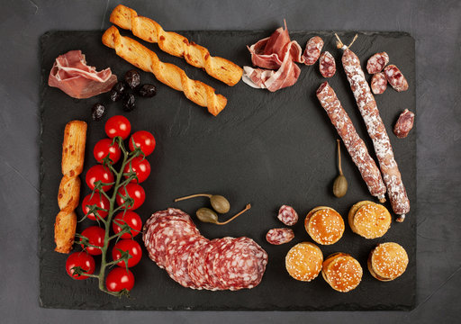 Appetizers table with differents antipasti, charcuterie, snacks and wine. Mini burgers, sausage, ham, tapas, olives, cheese variety board and baguette over grey concrete background. Top view, flat lay