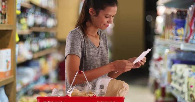 Pretty brunette female at supermarket with shopping list buying groceries, Young mother or wife with basket of items looking for what's missing, 4k