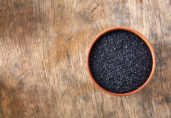 Black sesame seeds in a clay bowl on vintage wooden background, close-up, top view.