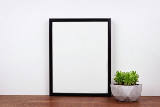 Mock up black frame with succulent plant on a shelf or desk. Wood shelf and white wall. Portrait frame orientation.