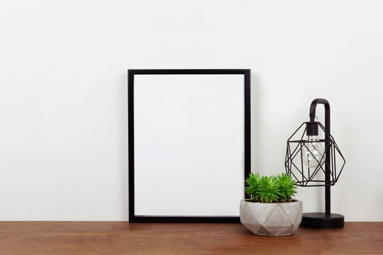 Mock up black frame, succulent plant and industrial style lamp on a shelf or desk. Wood shelf and white wall. Portrait frame orientation.
