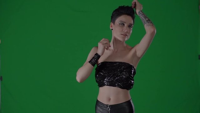 Young woman with short haircut is posing in photo studio. She is touching her clothing and body, looking at camera, chroma key green-screen