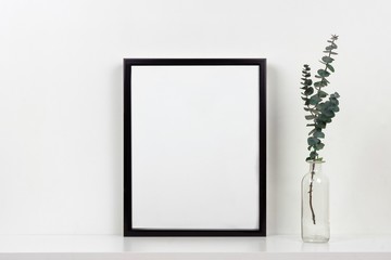 Mock up black frame with vase of branches on a shelf or desk. White shelf and wall. Portrait frame orientation.