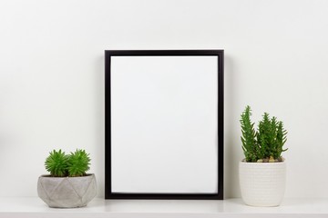 Mock up black frame and succulent plants in pot on a shelf or desk. White shelf and wall. Portrait...