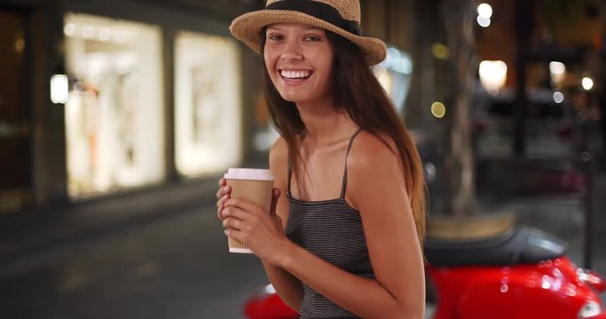 Smiling portrait of beautiful Caucasian girl in Italy enjoying coffee at night, Tourist woman wearing striped top and a fedora holding cup of coffee, 4k