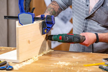 profession, carpentry, woodwork and people concept - carpenter with electric drill drilling wood...