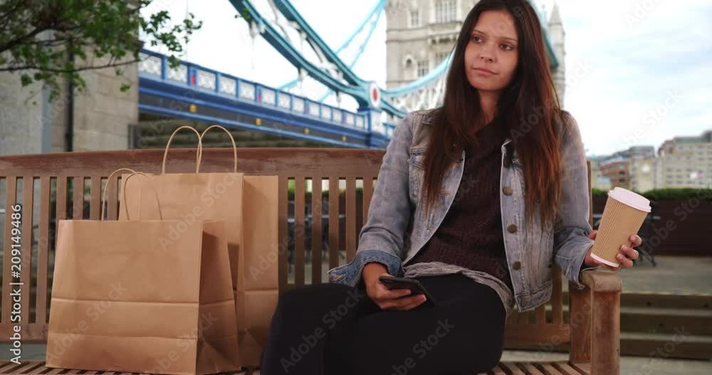 Wall mural hipster millennial female in jean jacket sits on bench by tower bridge with cup of coffee, trendy yo