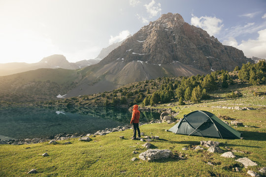 hiker in red jacket standing beside his tent in front of a mountain lake at surise