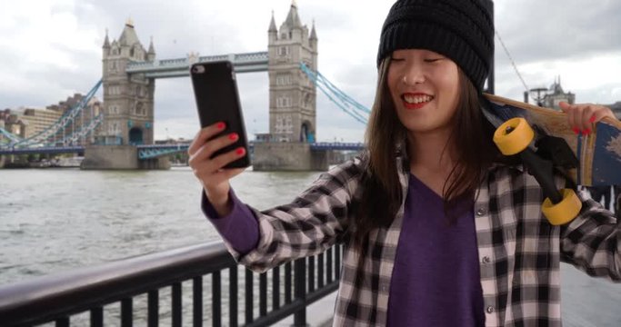 Happy hip millennial woman taking selfie with tech phone camera by Tower bridge in London UK, Close view of young Asian woman holding skateboard in Europe, 4k