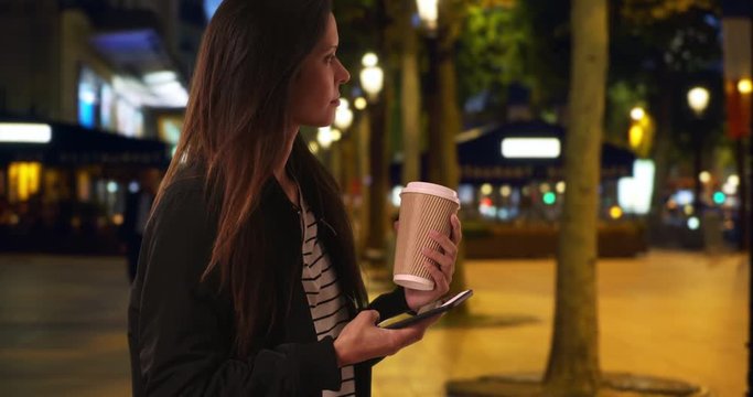 Hipster girl in bomber jacket with her coffee on the Champs-Elysees text messaging, Millennial female holding coffee while messaging friend with cell phone at night, 4k