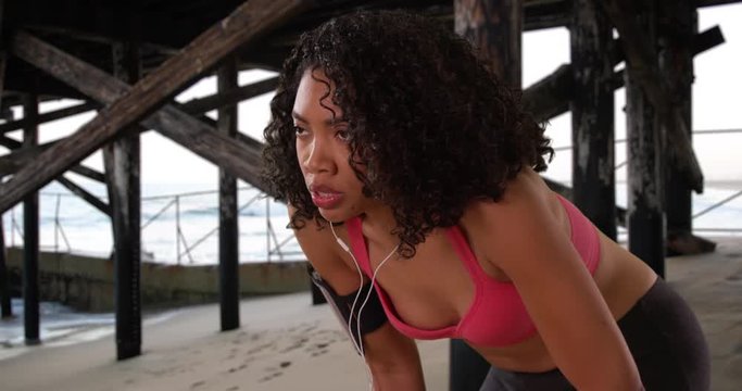 African American woman runner on the beach catching her breath and checking fitness tracker then running off screen, Black woman jogger resting under the pier with earbuds in then jogging away, 4k