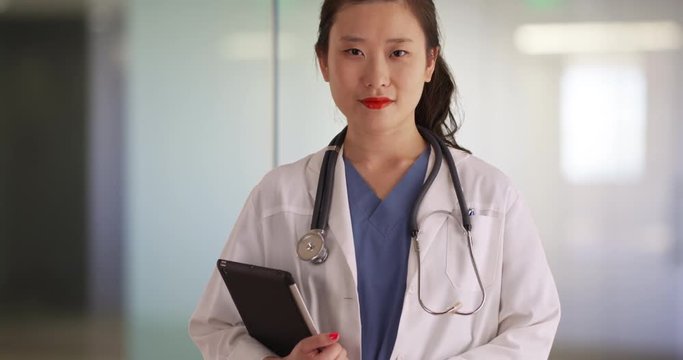 Professional female doctor carrying portable tablet computer and looking at camera inside empty office clinic, Young working medical practitioner holding tablet device indoors hospital hallway, 4k