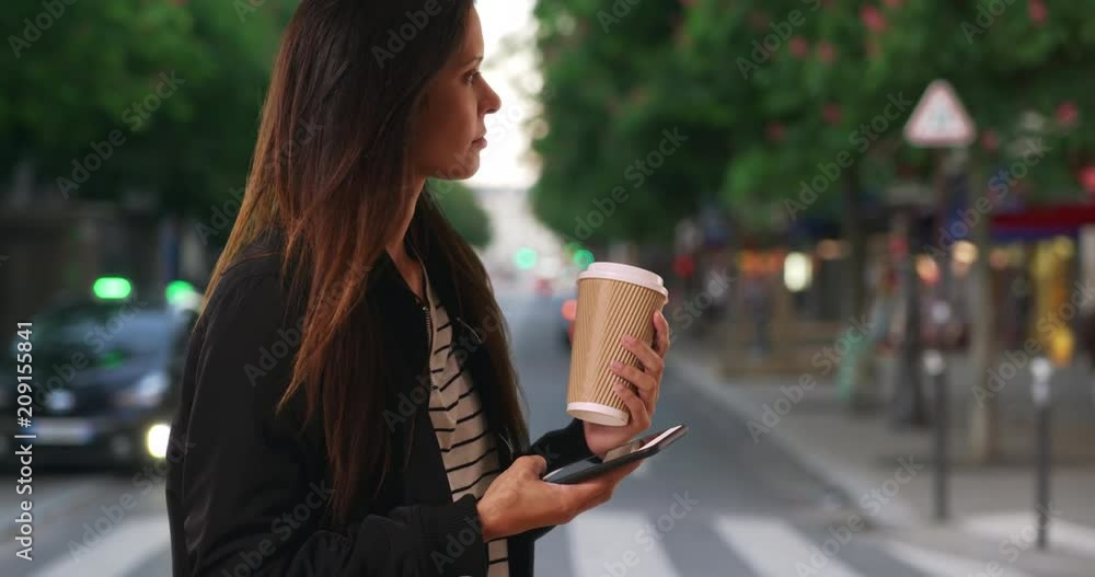 Wall mural Hipster girl in bomber jacket texting with cell phone while on city street, Millennial female with her coffee messaging a friend with mobile phone while standing outdoors, 4k