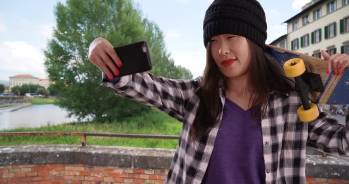 Cool millennial skater taking selfie with smartphone camera and holding skateboard in Florence Italy, Portrait of young Asian woman using technology to take picture in front of Arno river, 4k