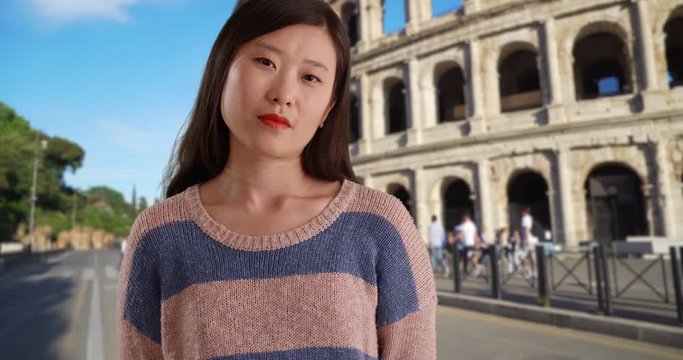 Serious young woman in static pose looking at camera in front of Colosseum in Rome, Close-up portrait of Asian millennial female with confident look in Italy, 4k