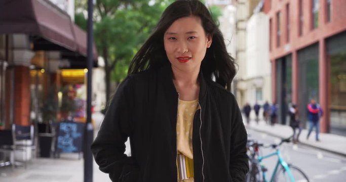 Millennial Chinese woman in black bomber jacket standing on city street with hands in pockets, Asian female stands on sidewalk with long hair blowing in the wind, 4k