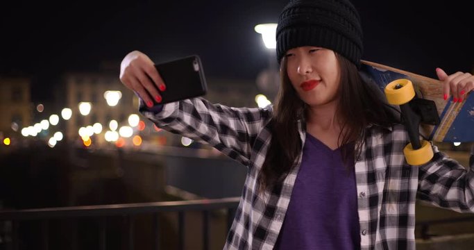 Cool millennial skater taking selfie with smartphone camera and holding skateboard on the Arno river in Florence Italy, Portrait of young Asian woman using technology to take picture in Europe, 4k