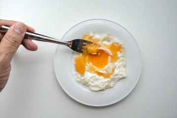 Hand holding fork to prick the egg yolk of beautiful fried egg in white plate isolated on white