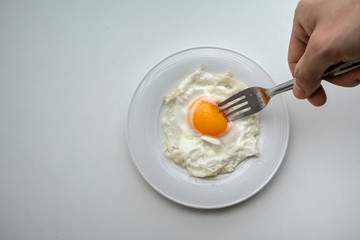 Hand holding fork to prick the egg yolk of beautiful fried egg in white plate isolated on white