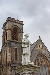 Fototapeta na wymiar Fort William, Scotland - June 11, 2012: Closeup of white stone war memorial statue on The Parade against gray sky. Shows young soldier in kilt contemplating, Duncansburgh Church of Scotland in back.
