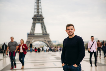 Cheerful man in dark blue pullover is standing on the background of the Eiffel Tower