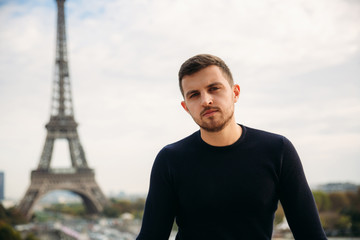 Handsome man wearing a dark blue pullover is standing on the background of the Eiffel Tower