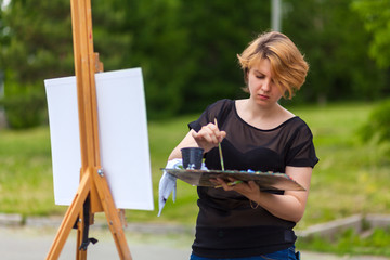 Artist  young woman paints on a canvas an urban landscape of a summer of oil paints in city park
