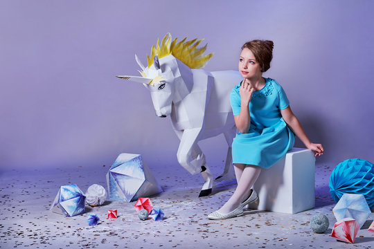 Dreamy,romantic little girl lady sits amid origami studio,white big unicorn,diamonds, geometric shapes. Beautiful,refined child in designer blue dress for holiday.Charming elegance teenager woman.
