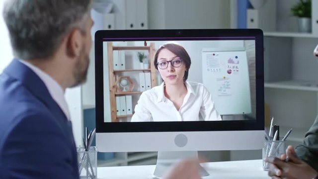 Tilt up of mid-aged caucasian businessman sitting at office desk with african colleague, waving and greeting businesswoman during video conferencing on computer