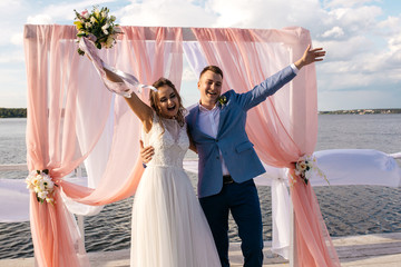 Happy bride and groom stand on the pier under the wedding arch