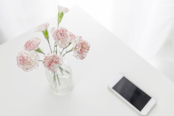 Top view of pink and white carnation flowers vase with smart phone on table in white room. Flat lay.