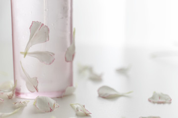 A Pink perfume bottle with pink and white carnation flowers and petals on white desk.