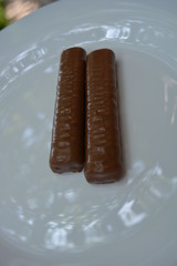 On a white plate of glass are two bars, a chocolate bar, shortbread cookies with caramel, covered with a layer of milk chocolate