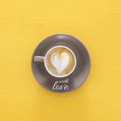 image of coffe cup with foam of heart shape over wooden yellow background and text: WITH LOVE.