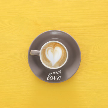 image of coffe cup with foam of heart shape over wooden yellow background and text: WITH LOVE.