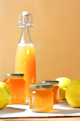 Homemade quince jelly and Juice in a bottle and glass jars with quinces on a linen tablecloth in bright sunshine in front of an yellow and orange background.