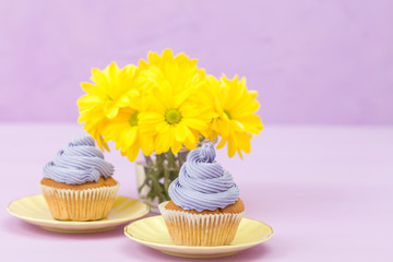 Cupcakes decorated with violet cream and bouquet of yellow chrysanthemums on violet pastel background with text area for greeting banner.